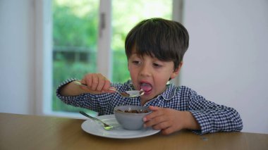 Mavi kareli gömlekli genç adam kahvaltılık gevrek yiyor, yeşil yapraklı arka planı pencereden görülebilen modern bir mutfakta yemeğine odaklanıyor.