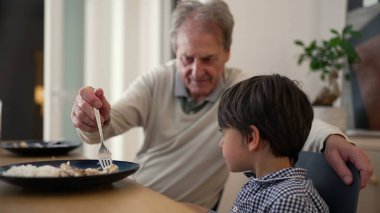 Yemek masasında kıdemli bir adam ve genç bir çocuk, birlikte yemek yemenin beslenme anına odaklanıyorlar. Sahne, bir ev ortamında aile bağlarını ve ilgili etkileşimleri vurgular