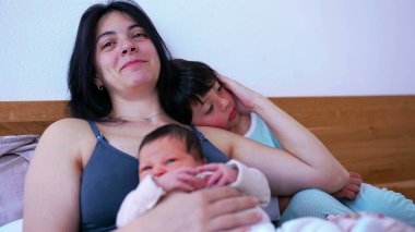 Gülümseyen anne, iki küçük çocuğu yataktayken rahatlar, bebeğini kollarında tutarken büyük çocuk omzuna yaslanır, sevgi ve sıcaklıkla dolu dokunaklı bir aile anını gösterir.