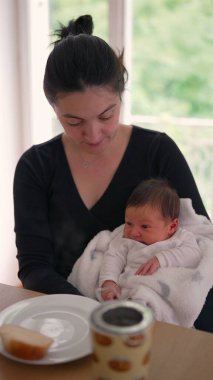 Anne bebeğini beyaz bir battaniyeye sarmış, bir parça ekmek ve kahve fincanıyla yemek masasında oturuyor, huzurlu bir anı paylaşıyor.
