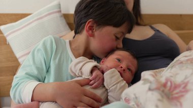 Büyük kardeş, anne kucağında dinlenirken bebeği nazikçe kucaklar. Kardeşler barışçıl bir anı paylaşıyor, samimi bir ev ortamında aralarındaki yakınlık ve sevgiyi vurguluyorlar. 