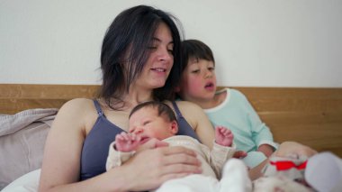 Anne, yeni doğmuş bebeğini kucağında tutarken yürekten bir hediye gösteriyor. Merakla izliyor, samimi bir aile anı. Sevgiye, kaynaşmaya, sıcaklığa ve ev yapımı hediyeye vurgu yapıyor.