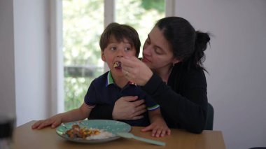 Anne oğlunu yemek masasında besliyor, besleyici bir aile anı yaşıyor. Çocuk yemek yerken şaşırmış görünüyor, yemek saatinde annenin gösterdiği ilgiyi ve ilgiyi yansıtıyor.