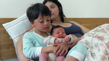 Büyük kardeş, anne izlerken bebeği kucağında tutuyor ve iç açıcı bir aile anı yaratıyor. Yaşlı çocuklar, bebeğe nazikçe dokunur ve ona bakar. Ailedeki yakınlık ve sevgiyi yansıtır. 