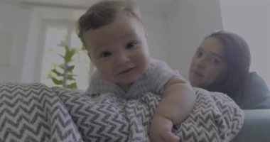 Bebek battaniyeye uzanıp duygularını ifade ederken anne, aydınlık ve rahat bir oturma odasında doğal ışıkla huzurlu bir aile ortamı yaratıyor.