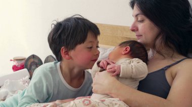 Anne bebeğini kucağında tutarken, büyük kardeş bakmak için eğilip birbirine bağlı aile bağını ön plana çıkarıyor. Sahne sıcaklığı, ilgiyi ve anne ile çocuk arasındaki sevgi bağını yansıtıyor.