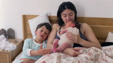 Anne bebeği tutarken, kardeş bebeğin ayağına hafifçe dokunuyor. Samimi aile anı, kardeşlerle anneleri arasında sıcak bir yuva ortamında paylaşılan sevgi ve bağın göstergesidir. 