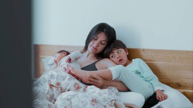Anne, bebeği ve büyük çocuğuyla birlikte yatakta, sessiz bir aile anını paylaşıyor. Bebek huzur içinde yatarken, büyük kardeş sevgi ve bağlantıyı yansıtıyor.