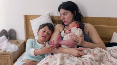 Anne bebeği tutarken, kardeş bebeğin ayağına hafifçe dokunuyor. Samimi aile anı, kardeşlerle anneleri arasında sıcak bir yuva ortamında paylaşılan sevgi ve bağın göstergesidir. 