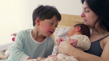 Anne bebeğini kucağında tutarken, büyük kardeş bakmak için eğilip birbirine bağlı aile bağını ön plana çıkarıyor. Sahne sıcaklığı, ilgiyi ve anne ile çocuk arasındaki sevgi bağını yansıtıyor.