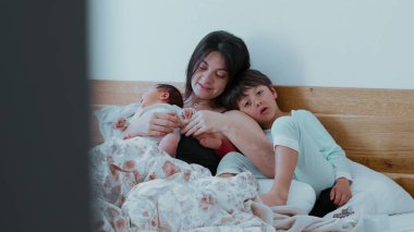 Anne, bebeği ve büyük çocuğuyla birlikte yatakta, sessiz bir aile anını paylaşıyor. Bebek huzur içinde yatarken, büyük kardeş sevgi ve bağlantıyı yansıtıyor.