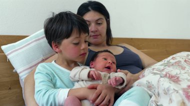 Older sibling holds the baby on their lap while mother watches, creating a heartwarming family moment. The older childs gentle touch and care for the baby reflect the close bond and affection within the family  clipart