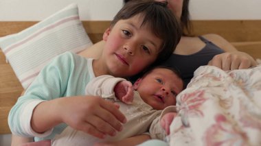 Büyük kardeş, anne kucağında dinlenirken bebeği nazikçe kucaklar. Kardeşler barışçıl bir anı paylaşıyor, samimi bir ev ortamında aralarındaki yakınlık ve sevgiyi vurguluyorlar. 