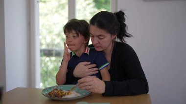 Anne oğlunu yemek masasında besliyor, besleyici bir aile anı yaşıyor. Çocuk yemek yerken şaşırmış görünüyor, yemek saatinde annenin gösterdiği ilgiyi ve ilgiyi yansıtıyor.