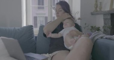Anne evde dizüstü bilgisayarla çalışırken bebeğini kucağında tutuyor, çocuk bakımını dengeliyor ve rahat bir oturma odasında uzaktan çalışıyor.