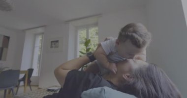 Anne, anne ve çocuk arasındaki bağ ve sevgi dolu doğal ışıkla dolu parlak bir oturma odasında oynarken, gülümserken ve gülerken bebeği havaya kaldırıyor.