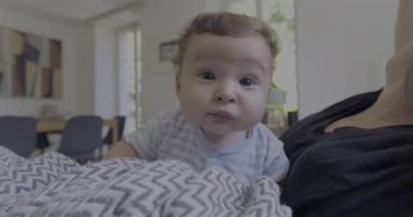 Bebek battaniyeye uzanmış merakla bakarken anne arka planda doğal ışık ve sıcak aile atmosferiyle dolu rahat bir oturma odasında gülümsüyor.