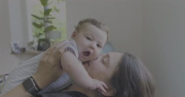 Anne, sevgi dolu ve sevgi dolu bir anda bebeği öpüyor ve kucaklıyor. Parlak bir oturma odasında oturuyor. Neşe ve yakınlık içinde kaynaşıyor.