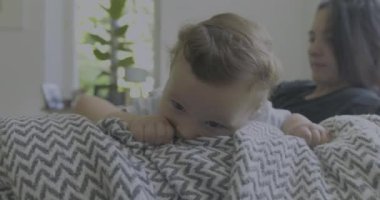 Bebek battaniyeye uzanmış, anne arka planda izlerken, keşfediyor ve oynuyor, sıcak aile anları, parlak ve rahat bir oturma odasında, bebeğin merakı...