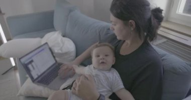 Anne kucağında bebek varken dizüstü bilgisayarla çalışıyor, iş ve ebeveynlik arasında denge kuruyor, rahat bir oturma odasında birden fazla iş yapıyor.