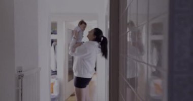 Annenin koridorda bebek kaldırma silueti, arka planda bulanıklık, ev içi, desenli duvar kiremitleri yansıması, samimi otantik yaşam tarzı, samimi aile sahnesi