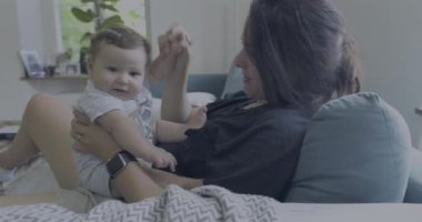 Anne ve bebek kanepede kaynaşıyor, bebek gülümsüyor ve meraklı görünüyor, sakin ve sıcak bir aile anının tadını çıkarıyor.