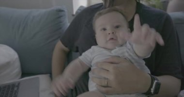 Anne kucağında bebekle evde dizüstü bilgisayarla çalışıyor. Kanepede birlikte sakin bir anın tadını çıkarıyor. Oturma odasını doğal ışık dolduruyor.