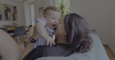 Anne sıcak ve sevgi dolu bir anda bebeği öpüyor ve kucaklıyor, doğal ışık sıcak bir oturma odasına akıyor, anne ve çocuk arasında neşeli ve samimi bir bağ kuruyor.