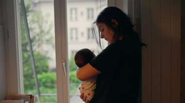 Anne yeni doğmuş bebeği pencerenin yanında tutuyor, huzurlu bir sahneye bakıyor, sıcak ve samimi bir anda sevgi, sevgi ve koruma hissi veriyor.