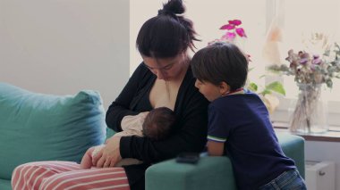 Anne, büyük oğlu izlerken yeni doğan bebeği koltukta emziriyor. Evde aile bağları, doğal ışık ve rahat, sevgi dolu bir atmosfer.