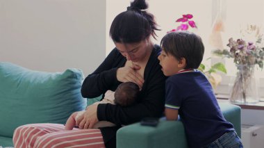 Anne, büyük oğlu izlerken yeni doğan bebeği koltukta emziriyor. Evde aile bağları, doğal ışık ve rahat, sevgi dolu bir atmosfer.