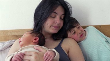 Anne bebeği kollarında tutarken, bileklikli büyük kardeş onun yanında oturup gözlerini ovuşturuyor. Sahne, aile hayatının hassas bir anını yakalar. Paylaştıkları bağı ve yakınlığı gösterir.