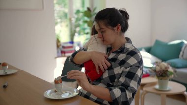 Anne, yeni doğmuş bebeği bir masada otururken kucağında tutuyor. Sakin bir evde anneliğin zorluklarını ve sevgisini gösteriyor.