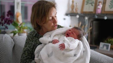 Büyükanne kundaklanmış yeni doğmuş bir bebeği kucağında tutuyor, şefkatle ve şefkatle aşağıya bakıyor, samimi bir oturma odasında yakınlık ve sevgiyi vurguluyor.