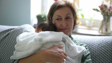 Yeni doğan torununu beyaz bir battaniyeye sarmış, sıcak, sevgi ve şefkat dolu bir bağ taşıyan, rahat, doğal aydınlık bir oturma odasında tutan yaşlı bir kadın.