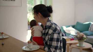Anne, yeni doğmuş bebeği bir masada otururken kucağında tutuyor. Sakin bir evde anneliğin zorluklarını ve sevgisini gösteriyor.