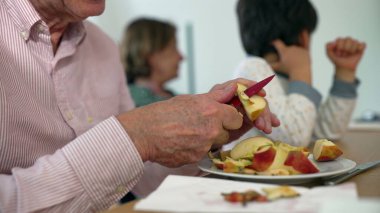 Elmayı bıçakla kesen kıdemli bir adam, arka planda izleyen küçük bir çocuk, ev içi aile ortamı, jenerasyon bağlantısını vurgulamak ve günlük basit görevler.