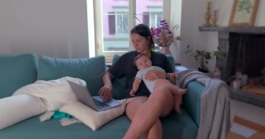 Anne kucağında bebeği kucağında tutarken rahat bir oturma odasında dizüstü bilgisayarla çalışıyor, işini ve aile zamanını aynı anda yönetiyor, görevlerine odaklanıyor.
