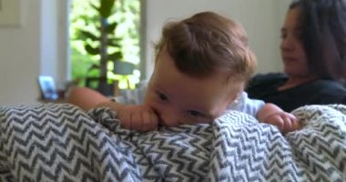 Bebek battaniyeye uzanmış, anne arka planda izlerken, keşfediyor ve oynuyor, sıcak aile anları, parlak ve rahat bir oturma odasında, bebeğin merakı...