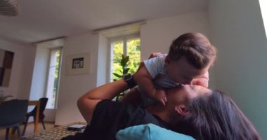 Anne, anne ve çocuk arasındaki bağ ve sevgi dolu doğal ışıkla dolu parlak bir oturma odasında oynarken, gülümserken ve gülerken bebeği havaya kaldırıyor.