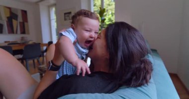 Anne sıcak ve sevgi dolu bir anda bebeği öpüyor ve kucaklıyor, doğal ışık sıcak bir oturma odasına akıyor, anne ve çocuk arasında neşeli ve samimi bir bağ kuruyor.