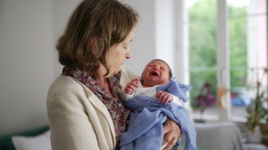 Mavi bir battaniyeye sarılı ağlayan bir bebeği kucaklayan ve rahatlatan yaşlı bir kadın, arka planda doğal ışık ve pencereyle kapalı bir yerde.