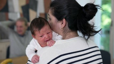 Bir annenin bebeğini omzunda tutarken, bebek huzur içinde dinlenirken, yaşlı bir adamın arka planda biraz bulanıklaşması, sıcak ve besleyici bir sahne yaratıyor.