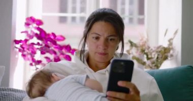 Anne kanepeye oturuyor, bebeğini kucağında tutuyor ve ev ortamında odaklanmış bir ifadeyle telefonuna bakıyor.