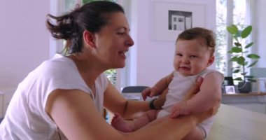 Anne sevgiyle bebeğini bir masada tutuyor. Parlak bir odada birbirine sevgiyle bakıyor. Şefkatli bir aile anını gösteriyor.