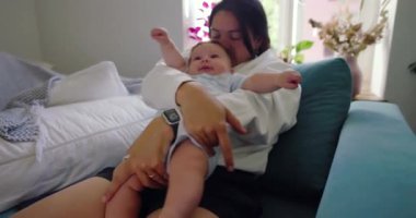 Anne kanepede bebeğe sarılıyor, bebek kucaklaşırken kolunu kaldırıyor, rahat ve sevgi dolu bir an yaratıyor.