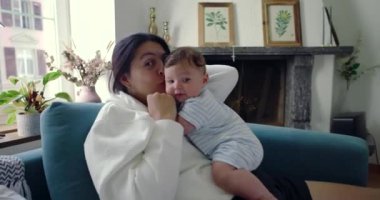 Anne bebeğe sarılıyor, bebeğin başını öpüyor. İkisi de rahat bir oturma odasında, kanepede huzurlu ve sevgi dolu bir anın tadını çıkarıyorlar.