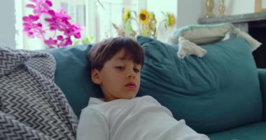 Rahat bir aile sahnesinde, bir anne bebeğiyle nazikçe ilgilenirken büyük oğlu, doğal ışığı olan parlak bir oturma odasındaki rahat bir kanepede uzanıyor.