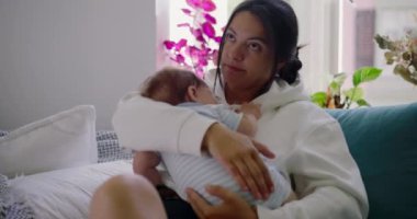 Anne, bebeğini nazikçe kucağına alıp bir kanepede kucaklar ve sıcak bir yuva ortamında sevgi ve özenle dolu huzurlu ve şefkatli bir an yaratır.