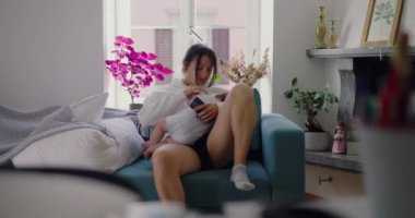 Anne kanepeye oturmuş bebeğini kollarına alıyor bebeğine tüm dikkatini veriyor. Huzurlu ve hassas bir an yaratıyor.